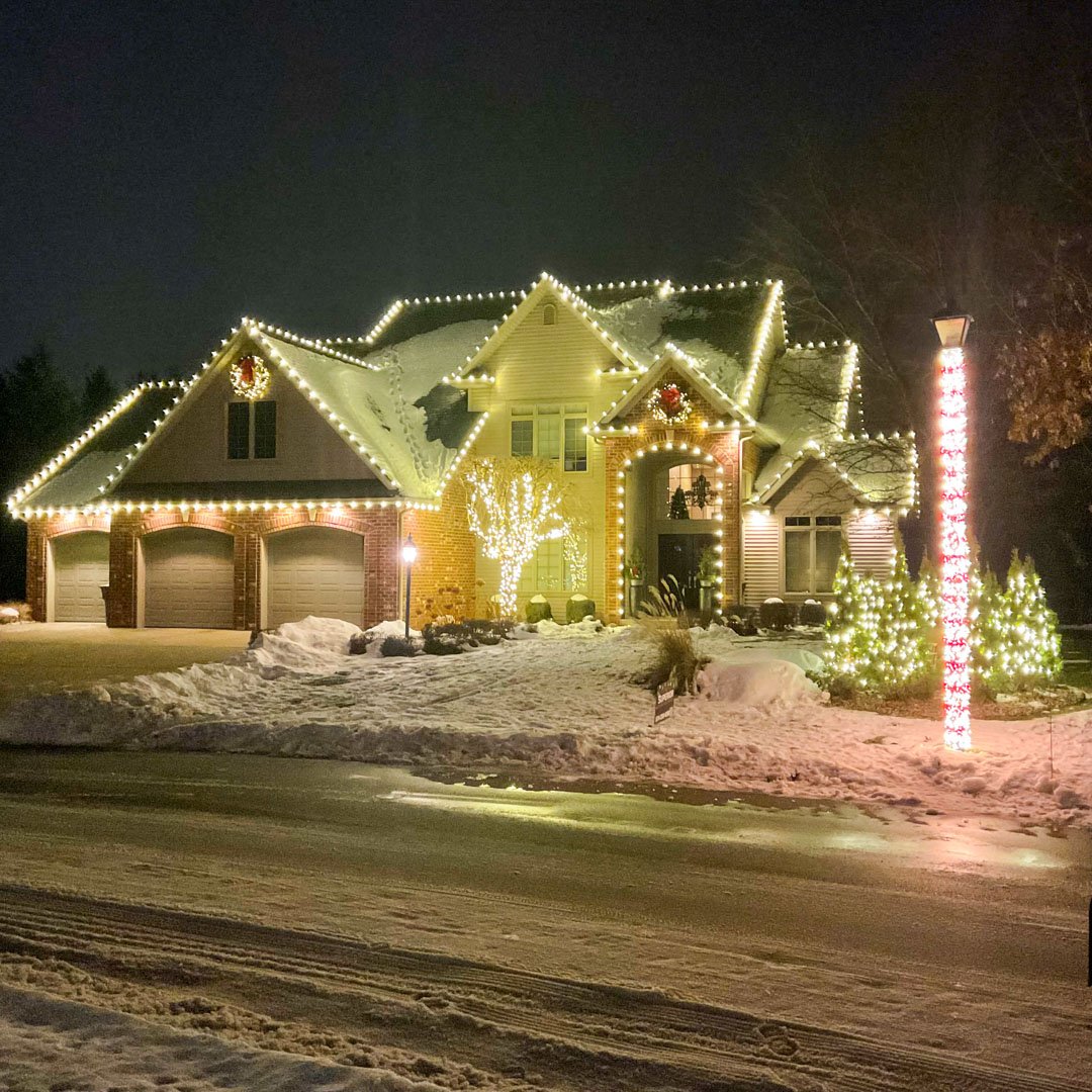 Warm white roofline lights, wreaths, and illuminated trees on a snowy home